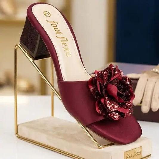 A single Foot Flexa heeled mule sandal in deep burgundy satin, featuring a large matching burgundy fabric and sequined flower embellishment on the toe strap. The shoe is displayed on a gold metal stand with a cream-colored base.