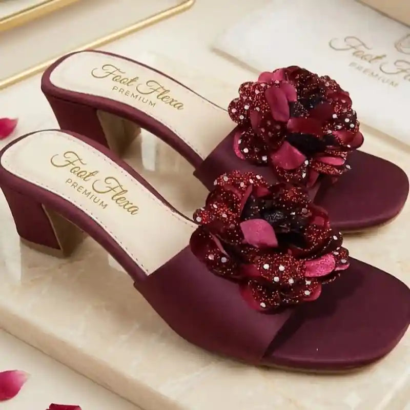 A pair of women's heeled mule sandals in deep burgundy satin, featuring large decorative floral embellishments on the straps made of matching burgundy fabric, glitter, and small clear rhinestones. The insoles are cream-colored with the brand name 'Feel Flexa PREMIUM' printed in gold script.