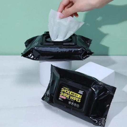 Black packaging of shoe shine wipes with a hand taking a wipe, on a light green background.