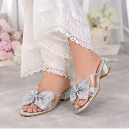 A pair of clear slide sandals with large silver glitter bows and low block heels, shown being worn with white lace-trim pants in a softly blurred indoor setting.