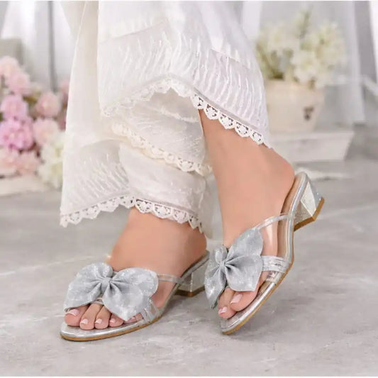 A pair of clear slide sandals with large silver glitter bows and low block heels, shown being worn with white lace-trim pants in a softly blurred indoor setting.