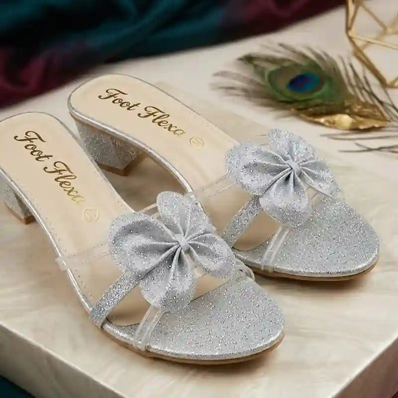 A pair of silver glitter mule sandals with sheer straps and large glittery bow embellishments on the toes, shown on a light wood surface with decorative peacock feather and fabric in the background. The insole displays the brand name 'Foot Flexa'.