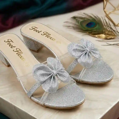 A pair of silver glitter mule sandals with sheer straps and large glittery bow embellishments on the toes, shown on a light wood surface with decorative peacock feather and fabric in the background. The insole displays the brand name 'Foot Flexa'.