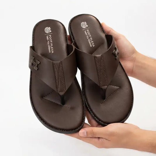 A pair of dark brown leather thong sandals with a prominent 'H' logo on the strap, held in two hands against a clean white background. The insole features the brand name 'FOOT FLEXA' and 'MADRID, SPAIN'.